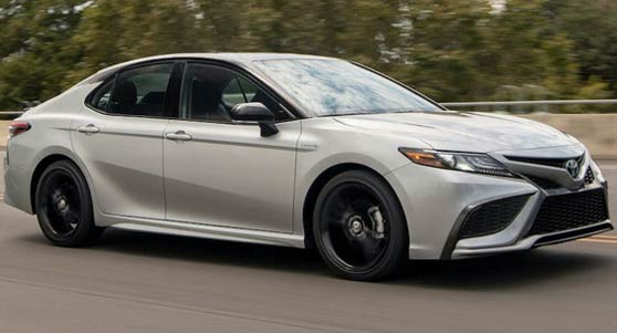 A picture of a 2023 Celestial Silver Metallic Toyota Camry LE driving on an elevated road with green tree tops sticking up from behind the guardrail in the background.