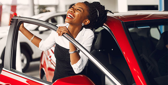 A woman is smiling and standing in an open doorway of a red car